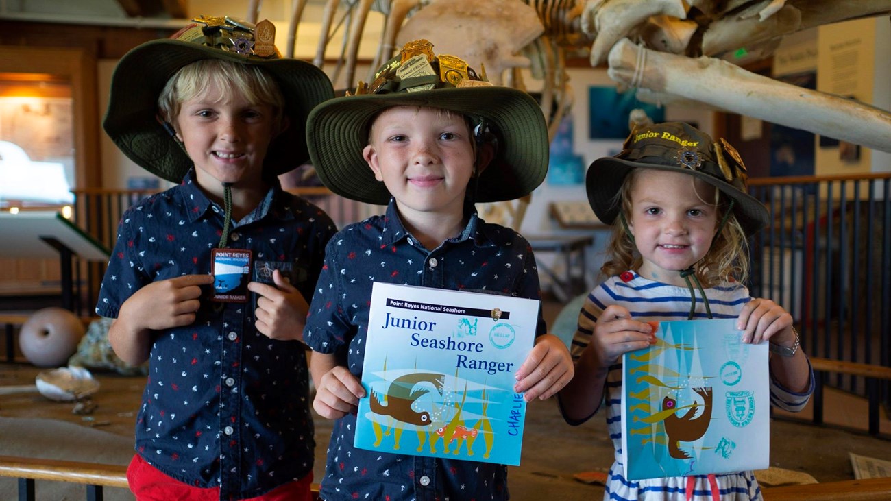 Three kids wearing ranger-like hats, smiling and holding up booklets in front of a whale skeleton