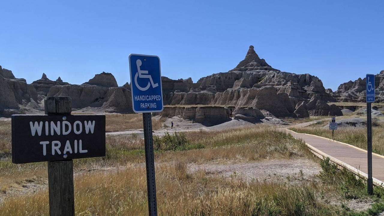 A sign marked Window Trail next to blue ADA sign with badlands buttes and boardwalk under blue sky.