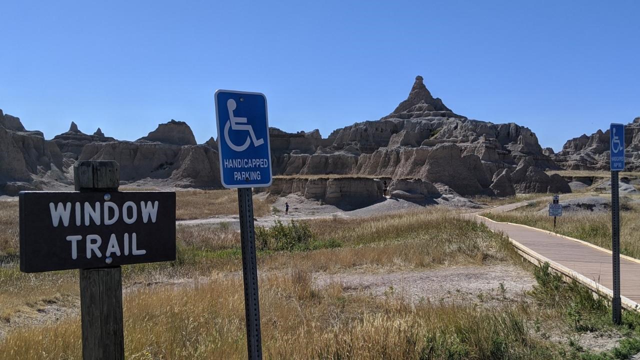 A sign marked Window Trail next to blue ADA sign with badlands buttes and boardwalk under blue sky.