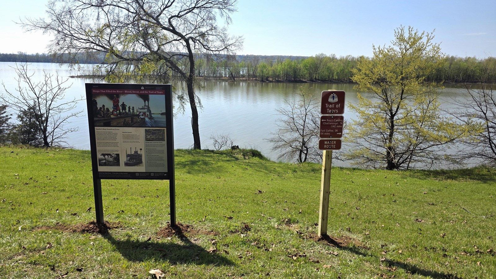 An upright wayside panel and three Trail of Tears route signs overlooking the Arkansas River.