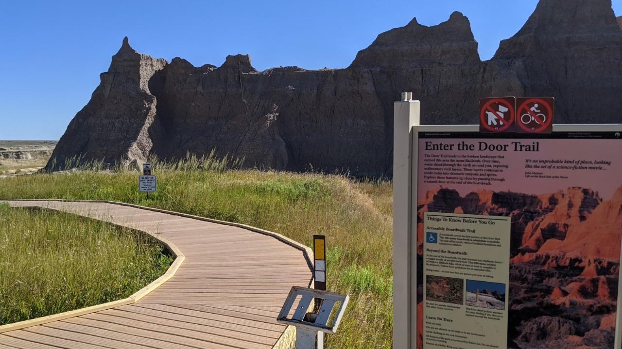 A sign marked Door Trail next to blue ADA sign with badlands buttes and boardwalk under blue sky.