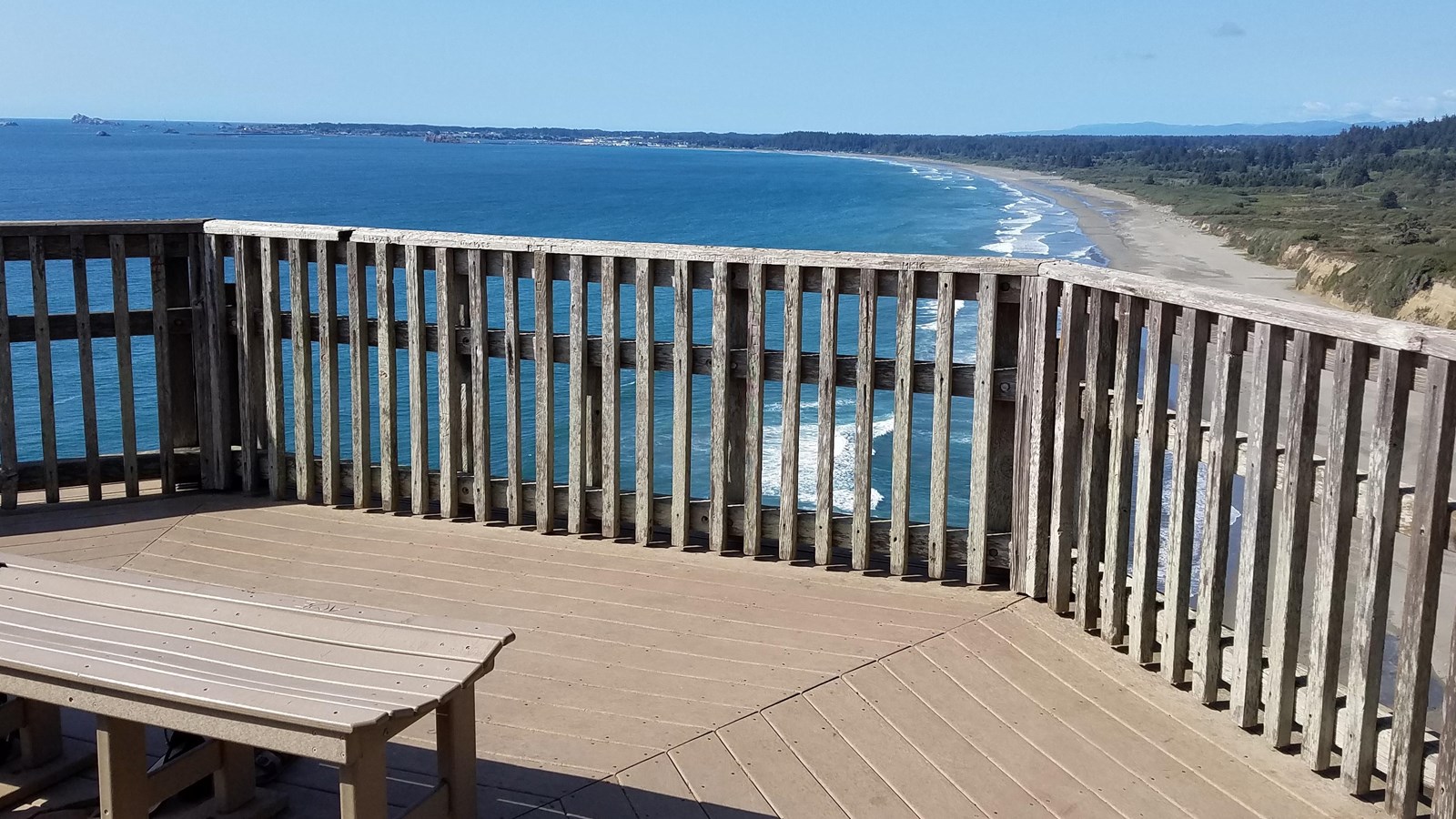 Wooden platform and handrails over look a blue ocean and sweeping beach.