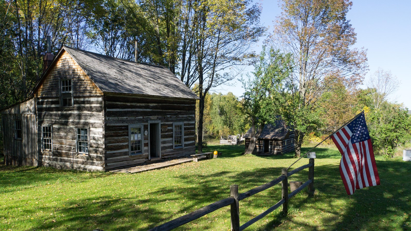 Summer scene of log cabins with a wooden fence in the foreground and trees in the background.