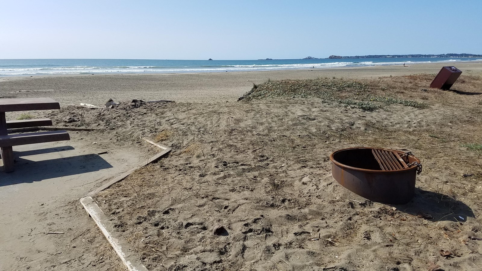 Fire ring and picnic table on a sandy beach. Blue waves break in the distance.