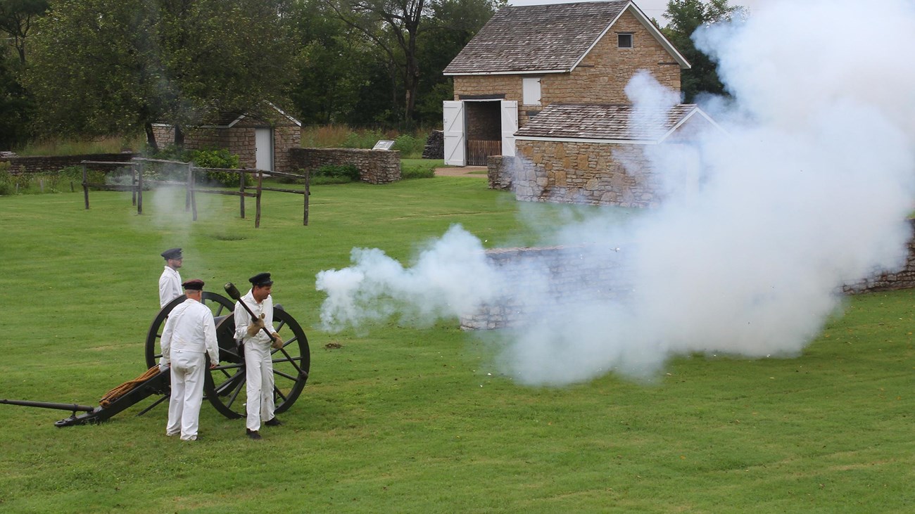 four men in white uniforms firing a cannon. Smoke is coming out of the cannon.