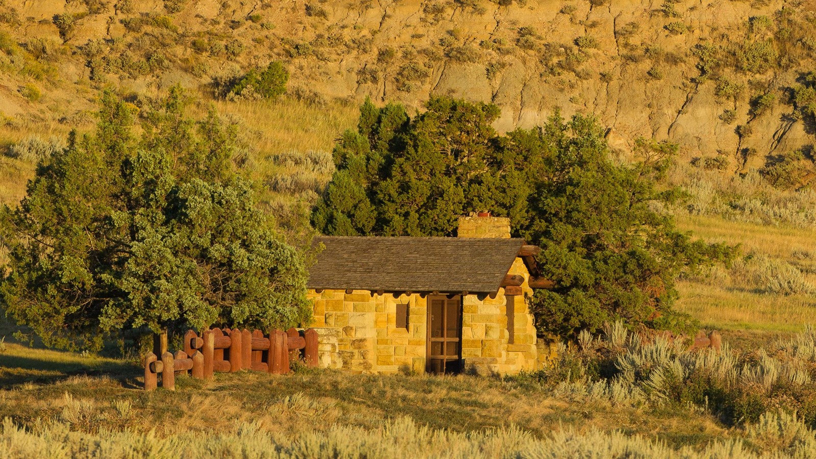 A small stone building sits at the base of a butte, surrounded by sagebrush with juniper behind it.