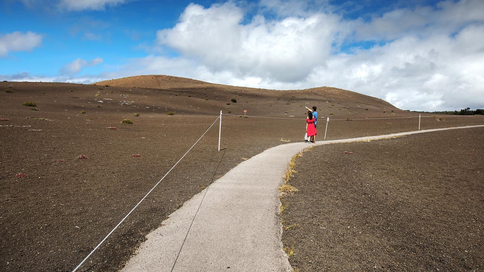 A man and woman walking on a paved path through a barren landscape covered by cinders