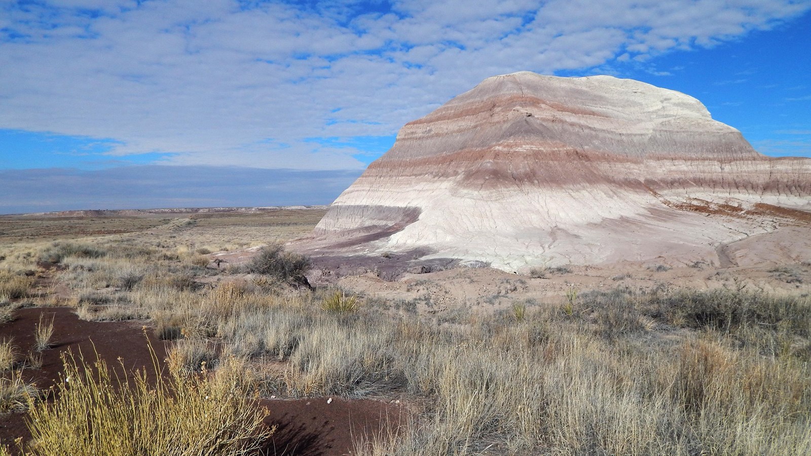 Old road winding through grassland in front of banded badlands under a blue sky with some clouds