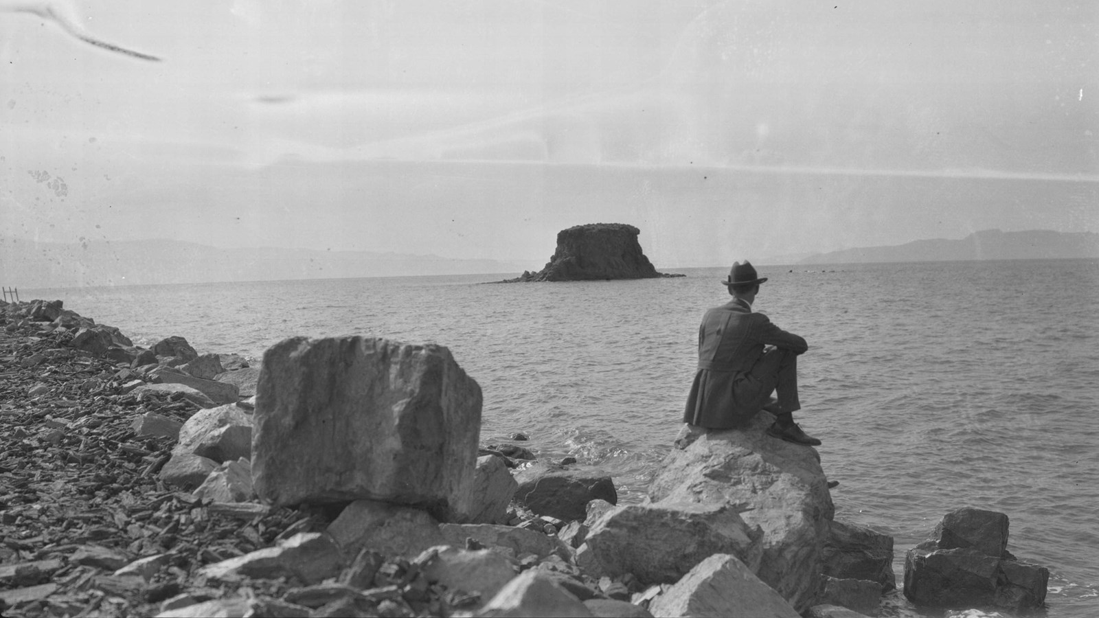 Black and white, man sits on rock at edge of Great Salt Lake. Black Rock is small in the distance.