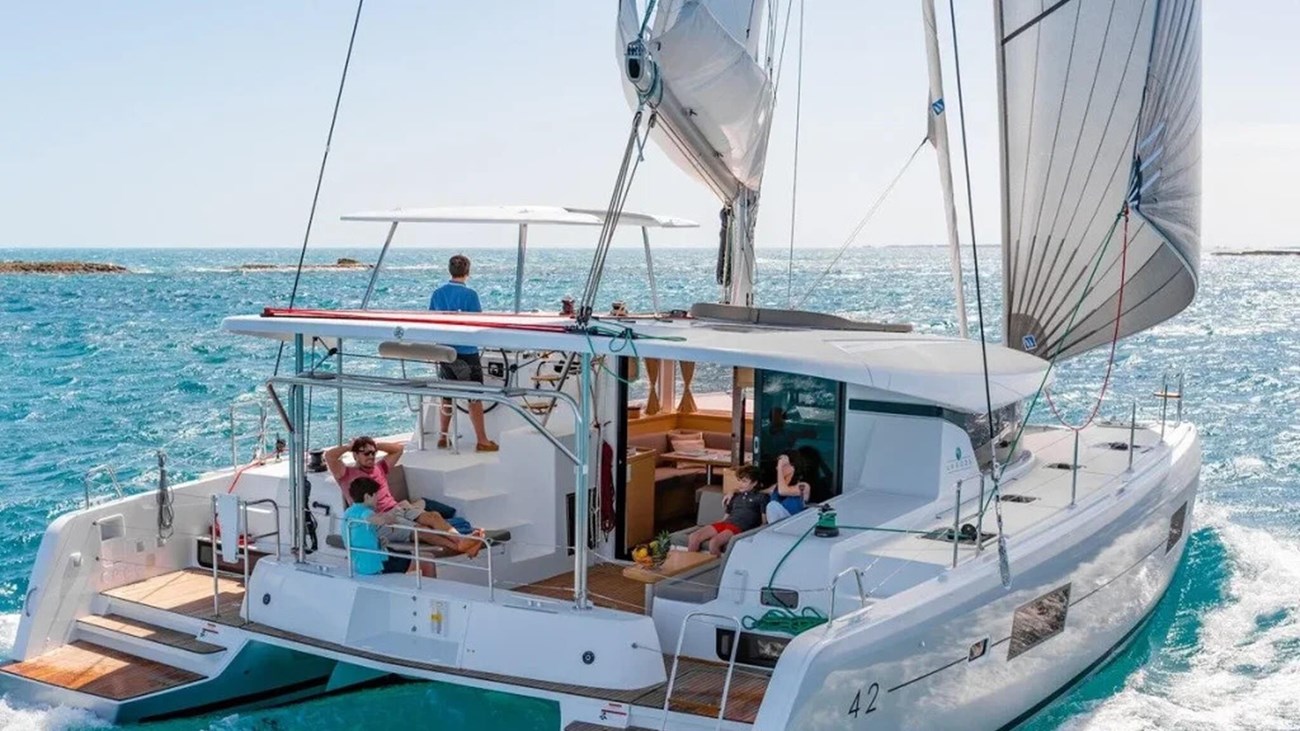 A catamaran with sails up, hosting people on deck, sails on a bright blue ocean.