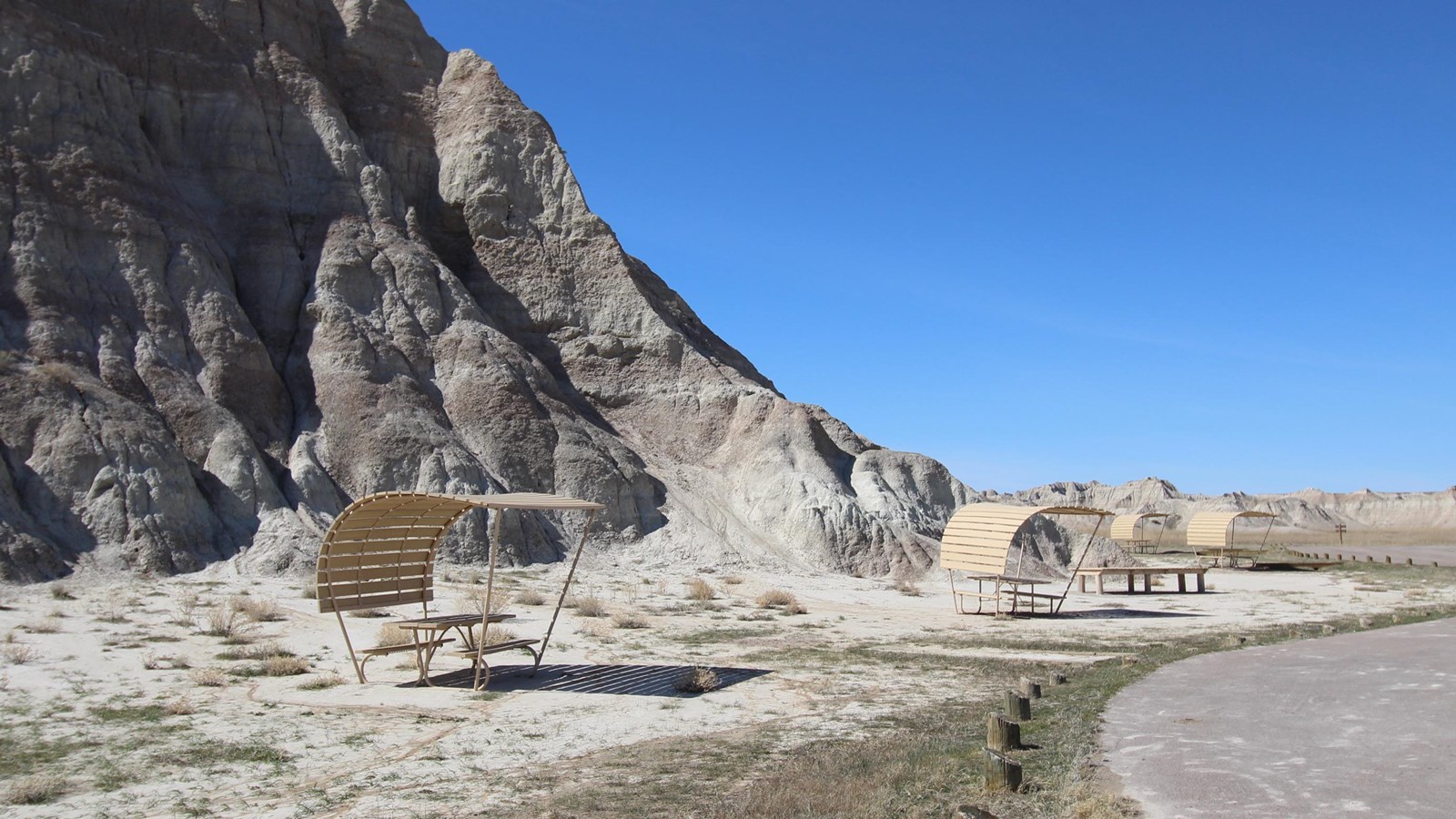 Two shaded picnic tables beside paved road, badlands butte, and under a blue sky.