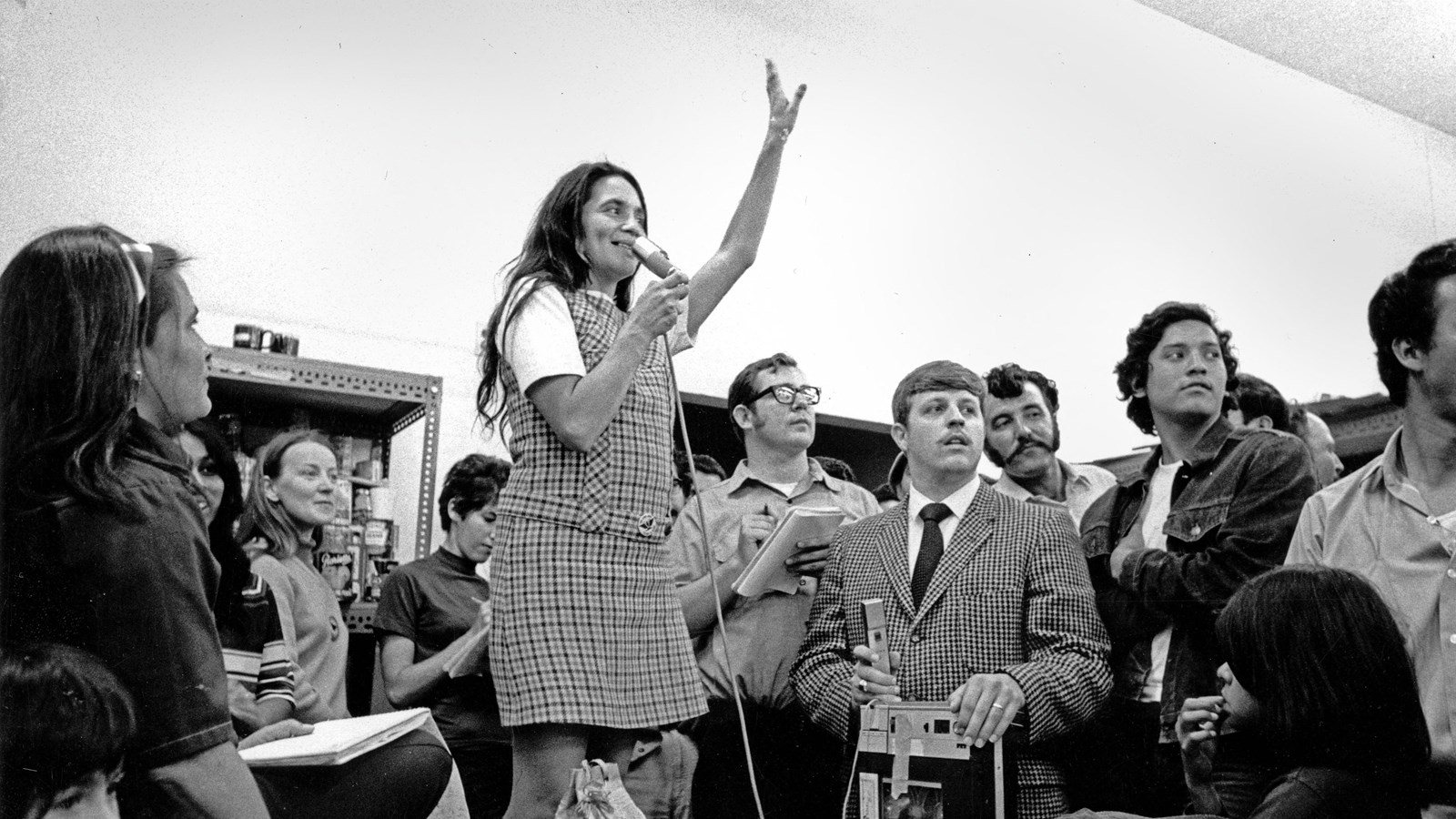A woman stands on a chair, arm raised, speaking into a microphone while surrounded by people