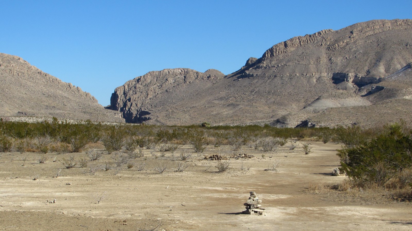 A dirt trail leads across the flat desert to a rocky wash, which bisects a mountain ridge. 