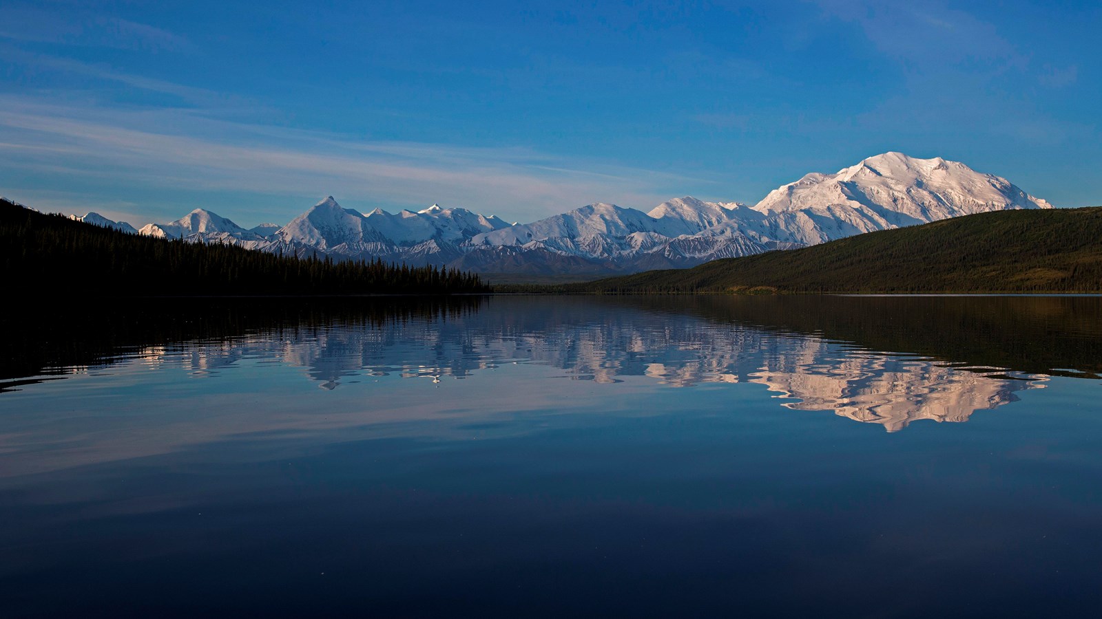 A long blue lake with Denali and other snowy mountains in the distance.