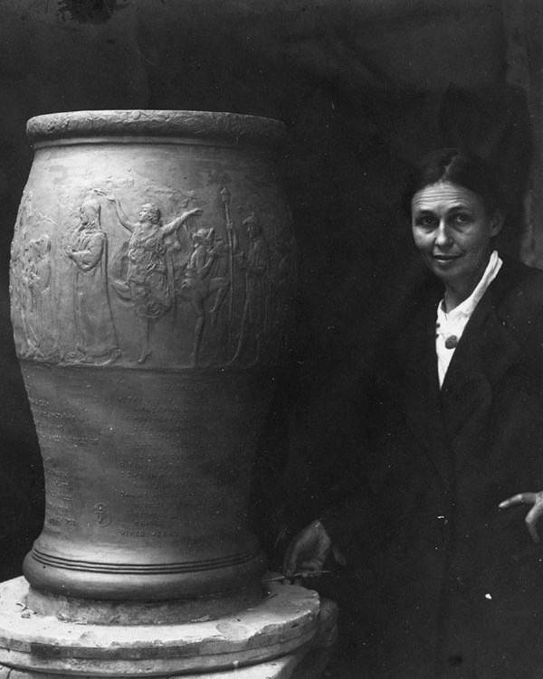 Woman in long black coat with hand on hip next to large urn with lettering and Greek figures.