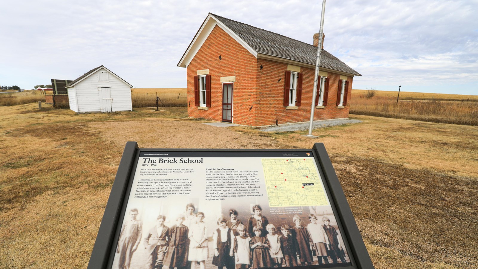 NPS wayside is situated in front of one room brick schoolhouse in a prairie setting.