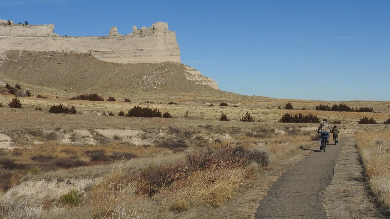 A pair of bicyclists are seen riding towards a distinctive sandstone rock formation. 
