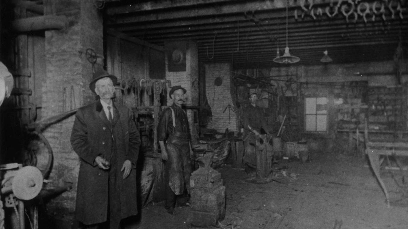 A man in a suit and coat stands in the foreground of a blacksmith shop floor.