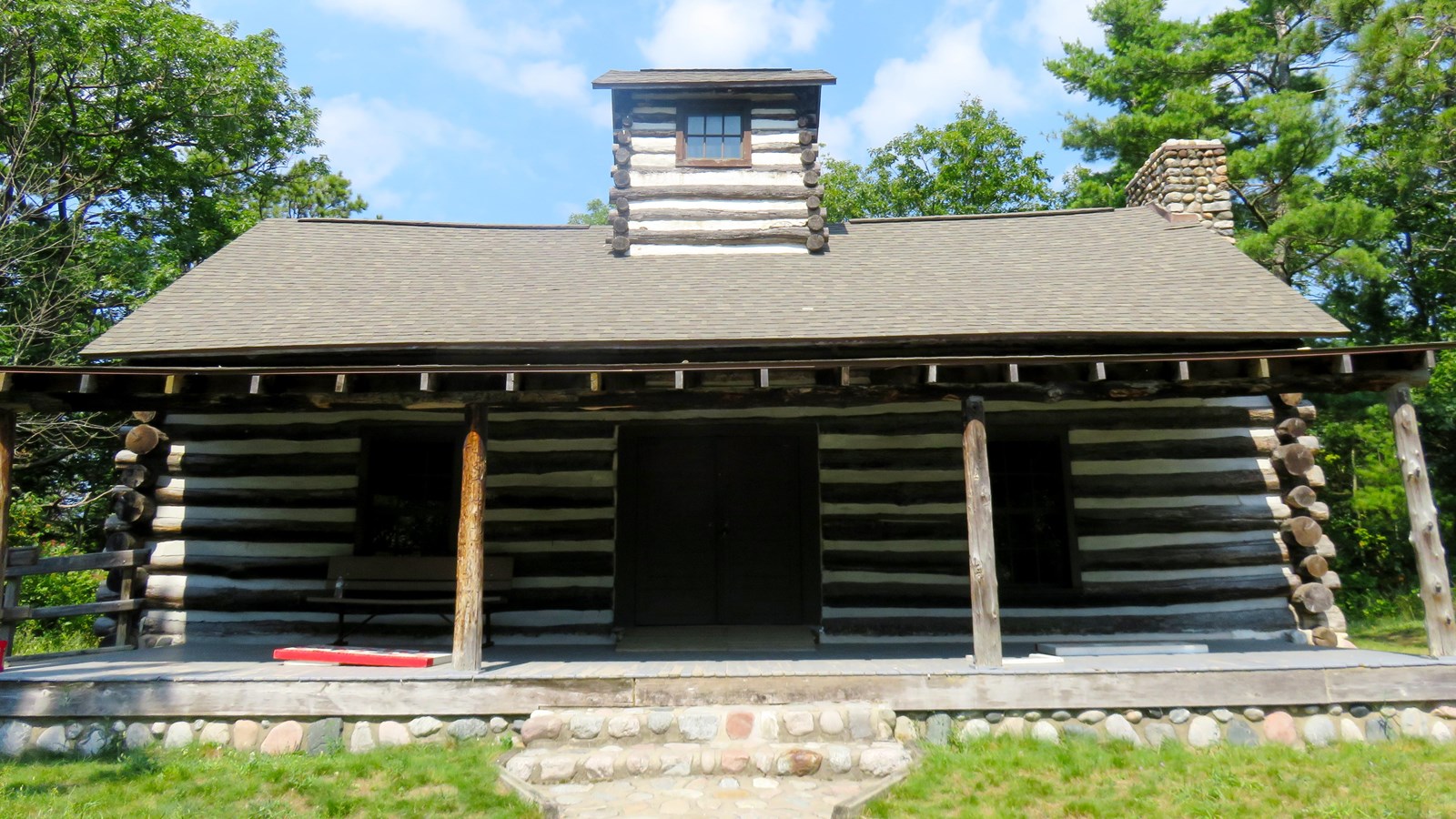 Log cabin with dark brown logs and white mortar and wide covered porch sits on a stone foundation