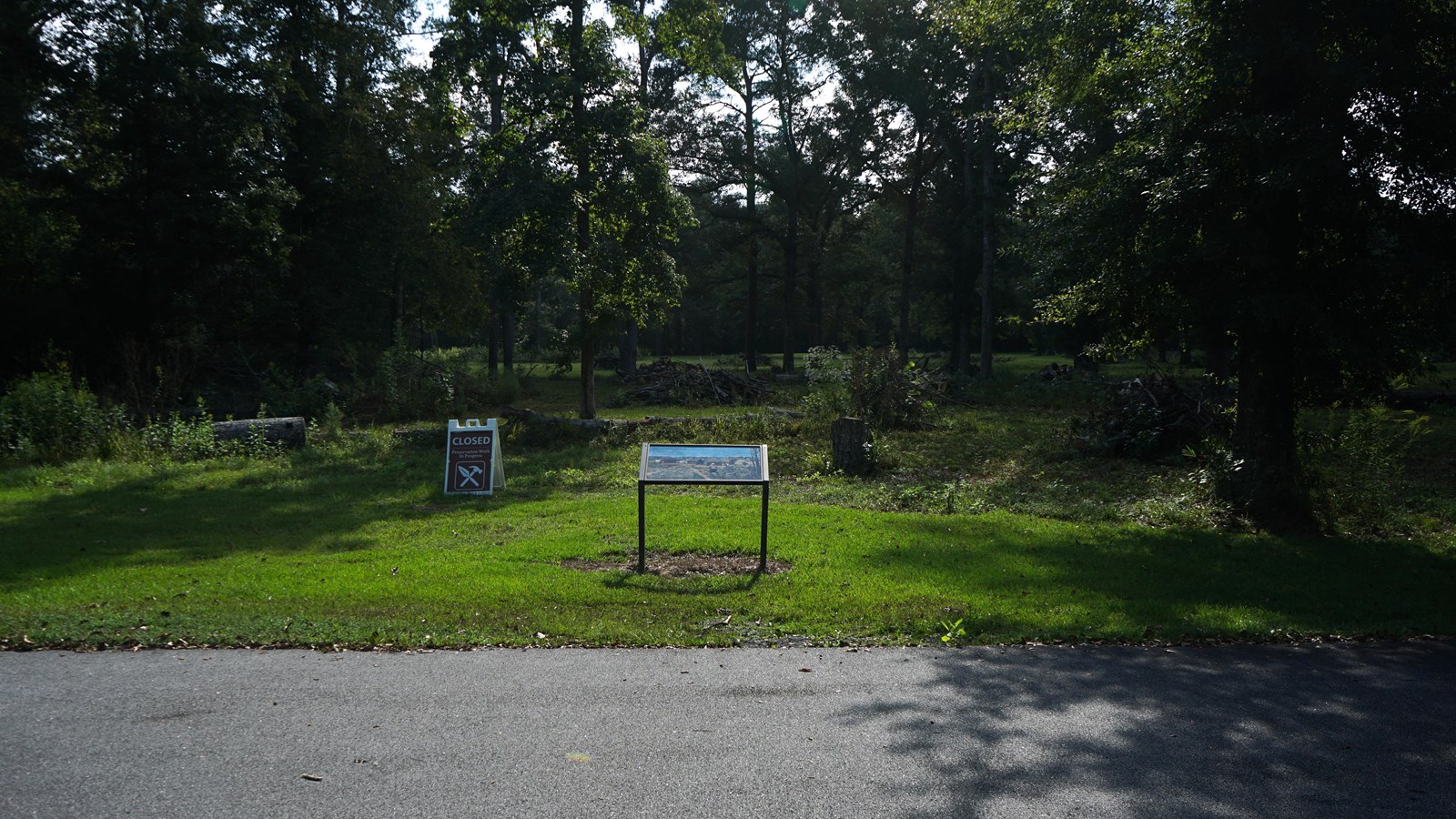 The image displays a metal informational wayside next to a roadway surrounded by forest.
