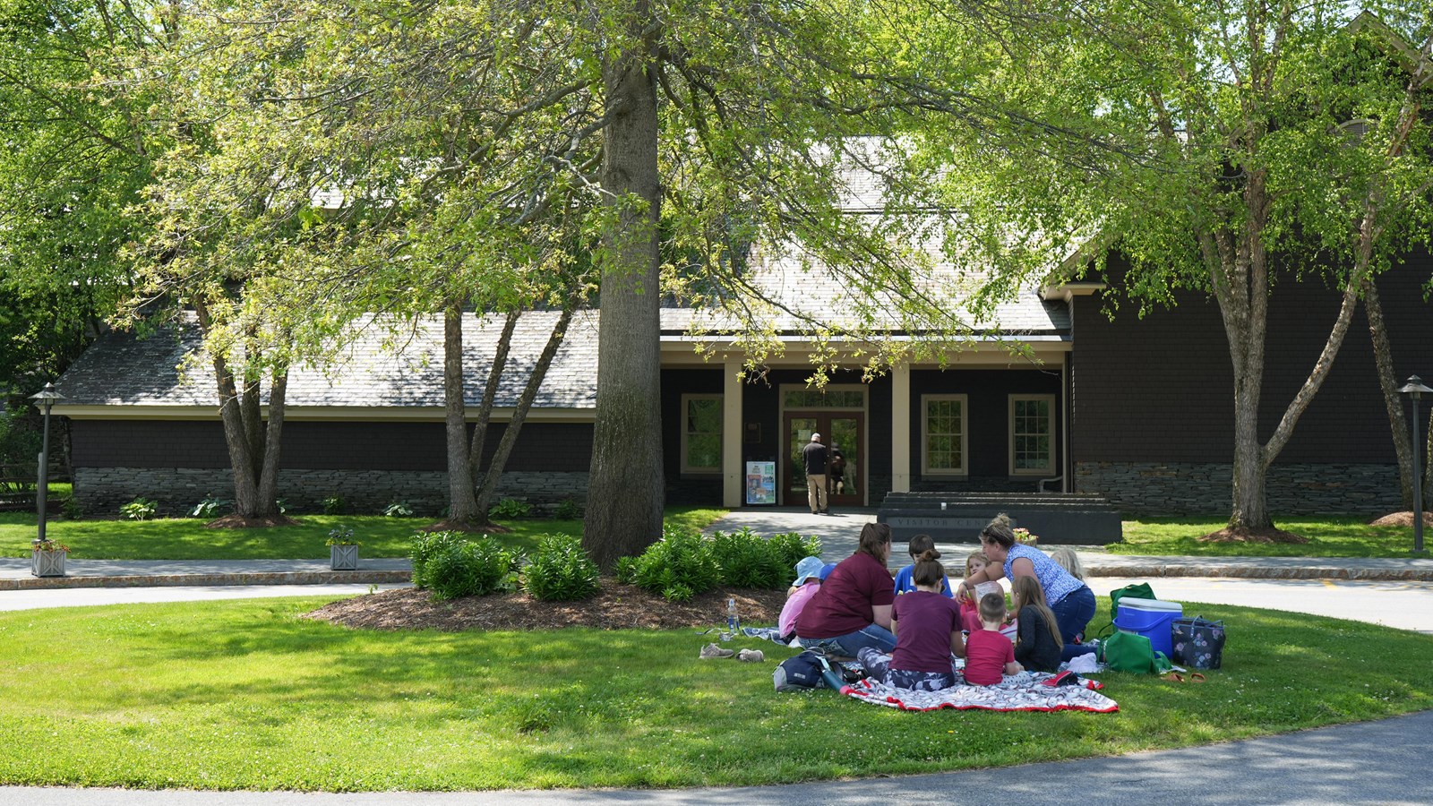 dark brown building with light brown roof in background with large tree and 10 people on blanket