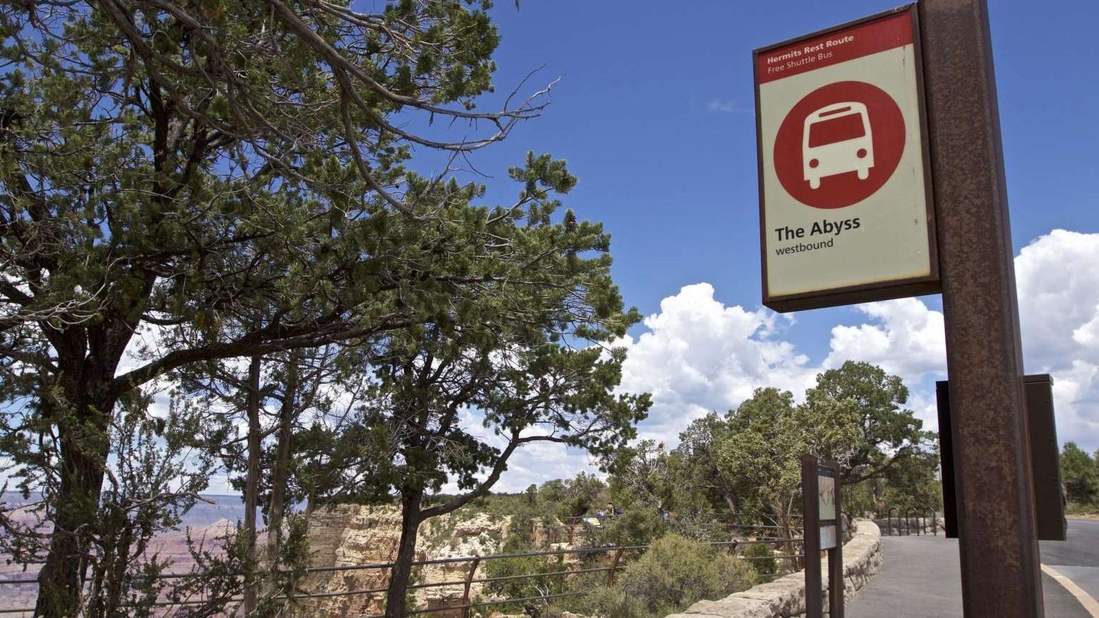 A low rock wall separates a bus stop, marked by a red and white sign, from the rim of Grand Canyon.