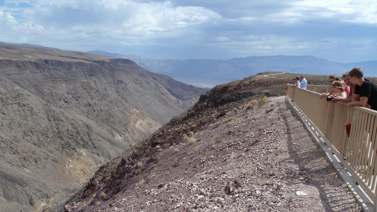 Visitors lean over a metal fence to look into a colorful deep desert canyon with dark clouds above.