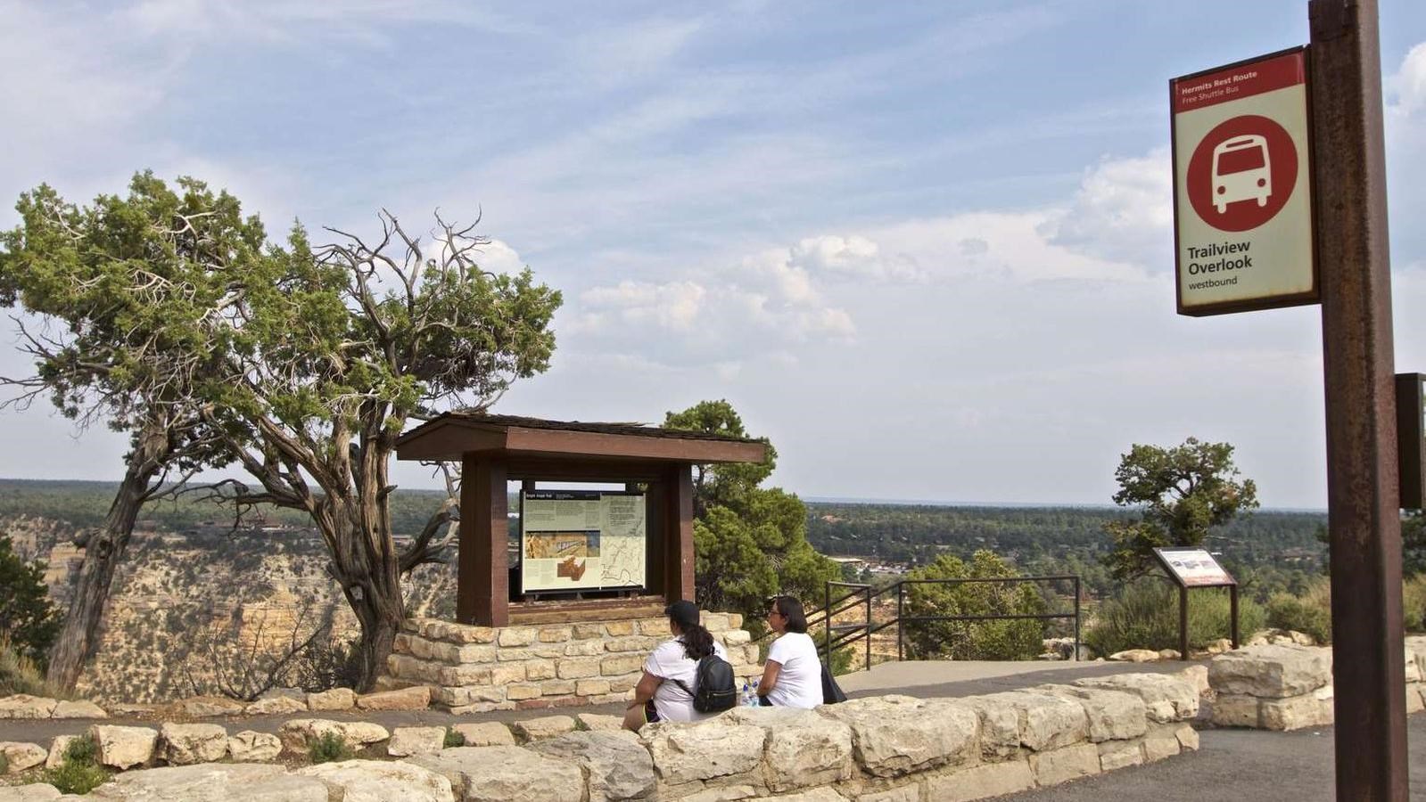 A couple sits on a rock wall along the rim of Grand Canyon next to a shuttle stop with a red sign.