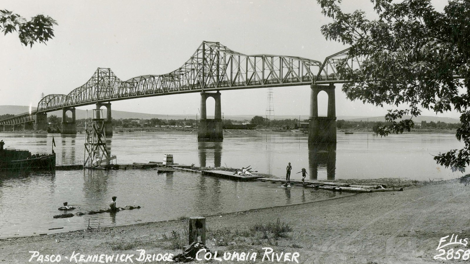 A black and white photo of a bridge spanning a large river. There are people playing in the river.