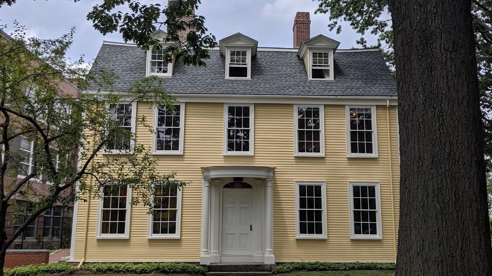 Yellow two-story house with large windows, white front door, gray roof, and red chimneys.