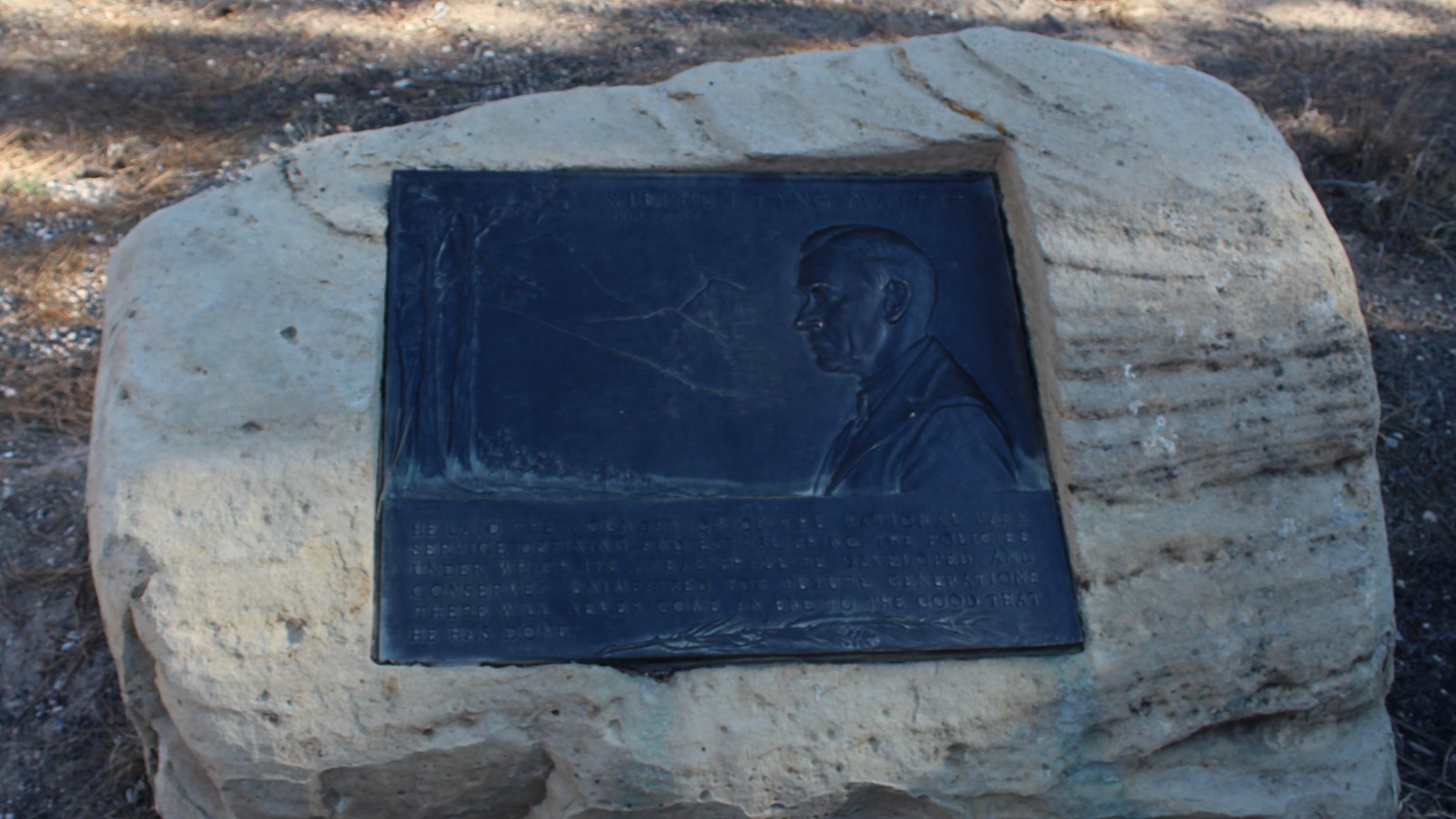 A dark metal plaque with raised relief profile of a man and text embedded in a large boulder