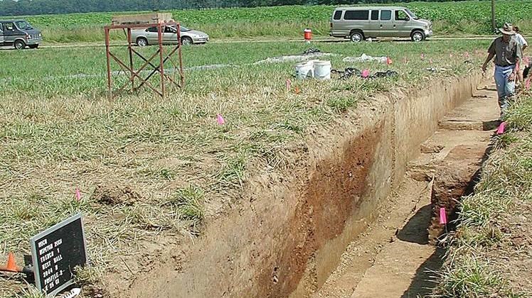 An archeological excavation trench shows red soil piled on yellow soil.