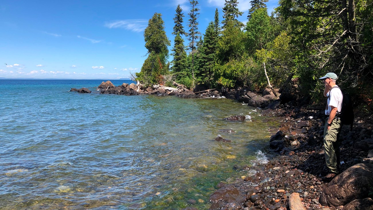 A person stands on a rocky beach next to a lake with a forest in the background. 