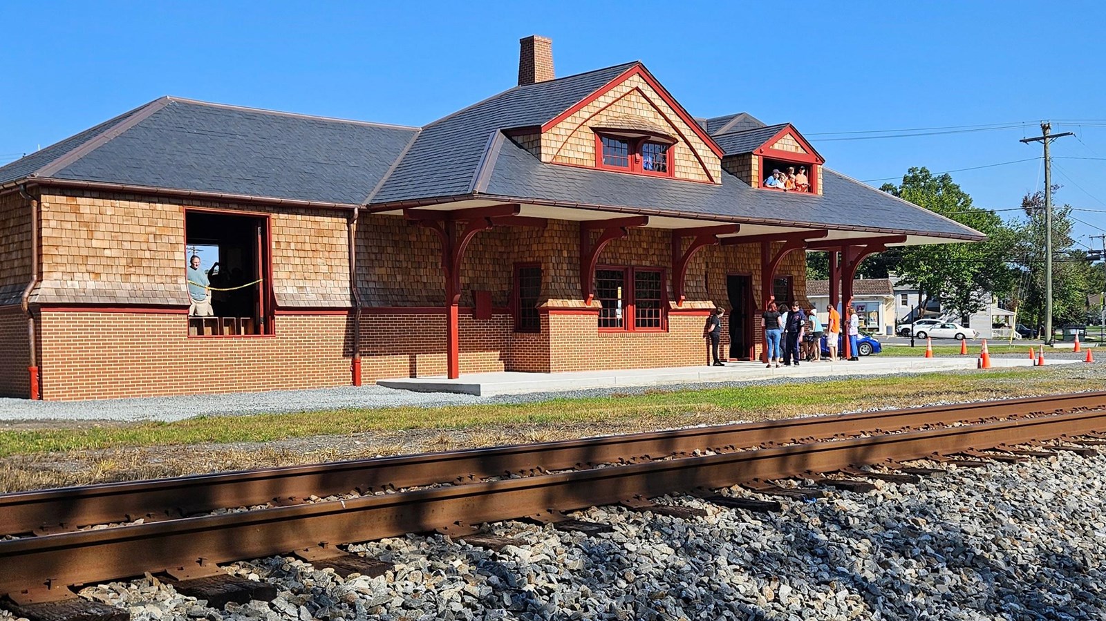 An historic brick and wood train station with slate-gray roof and a group of people on the platform.