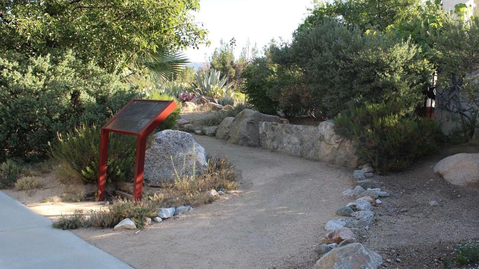 A dirt path lined with rocks. Next to the trail red metal posts hold a dark colored sign.
