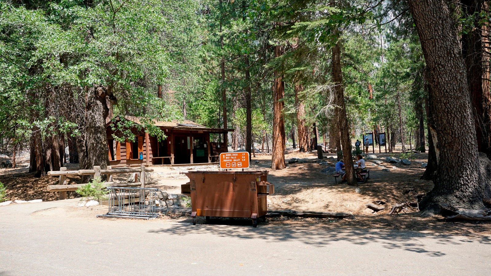 A small brown building sits in the shade of tall trees. A dumpster stands in front of it