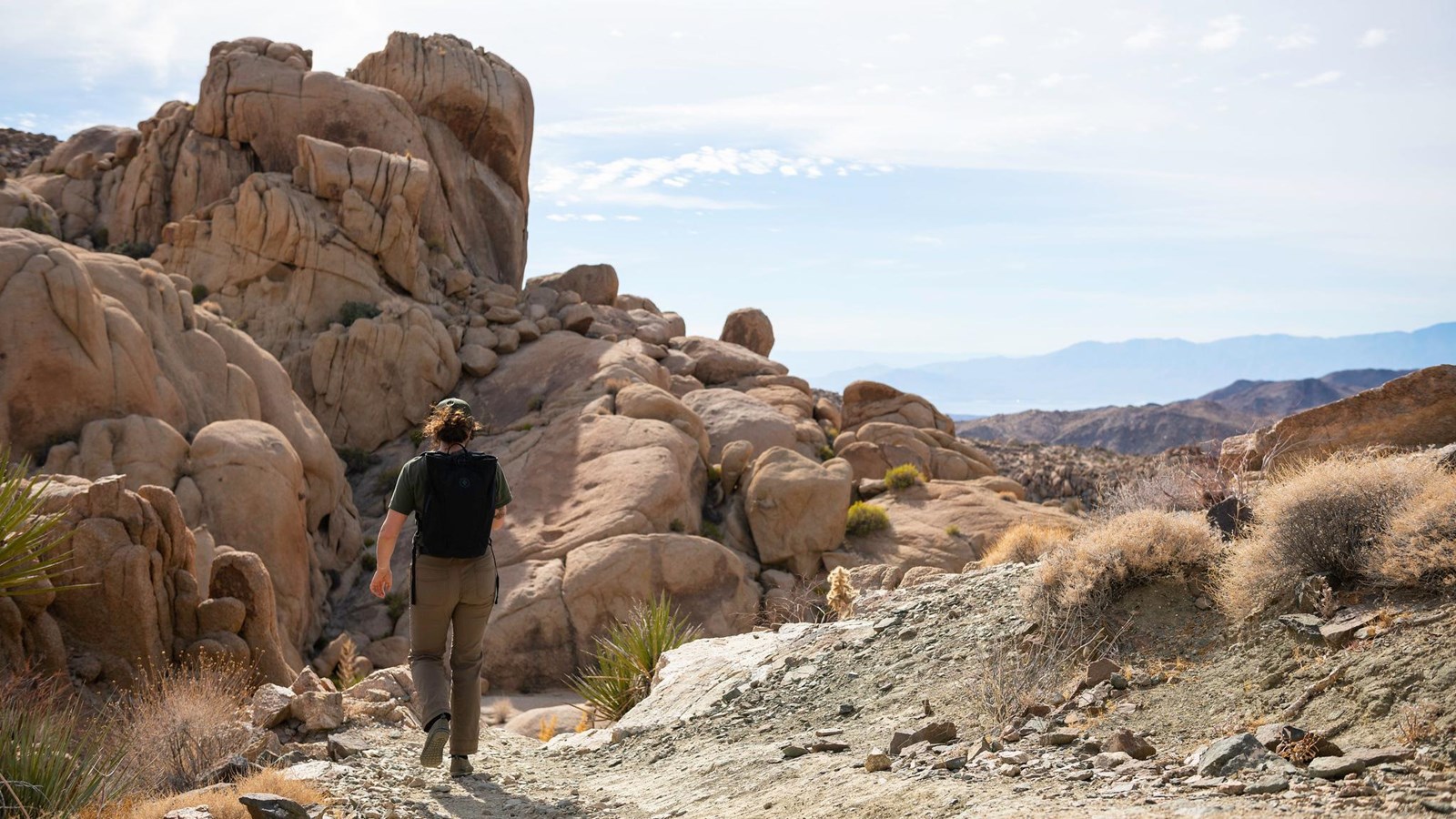 A hiker walks down a scree-covered trail next to tall boulders.