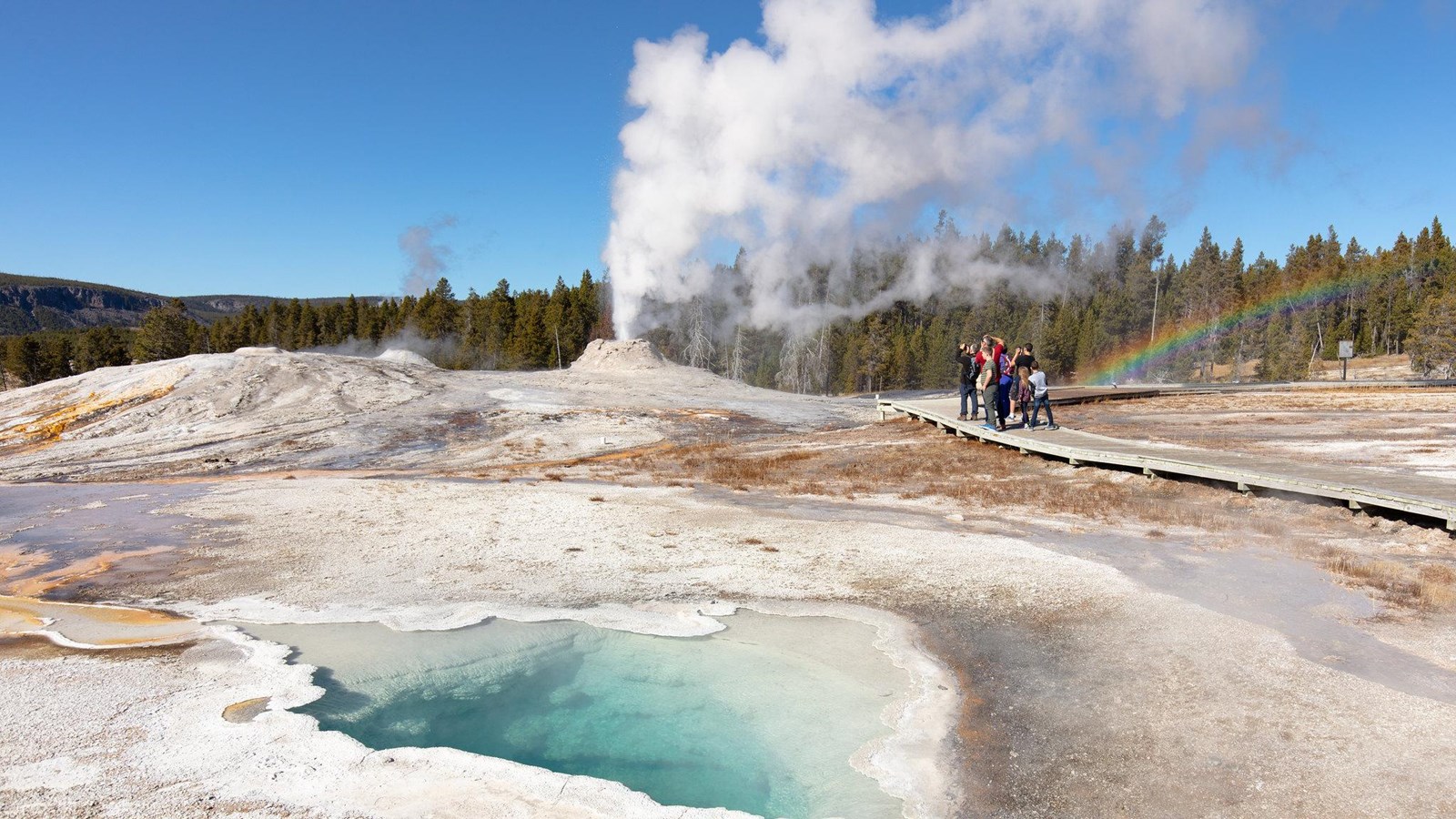 People walk along a boardwalk as water shoots into the air from a geyser cone and a rainbow appears.