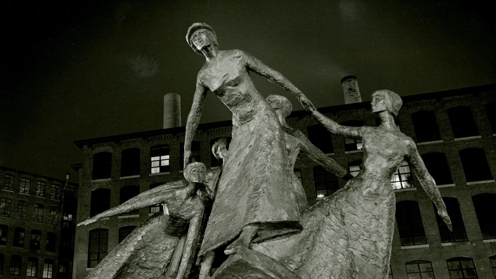 A bronze sculpture of five women in front of a historic mill building