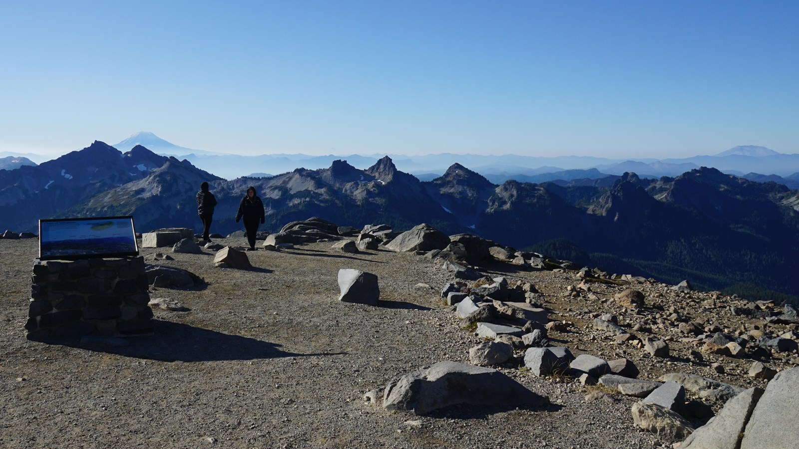 A wayside panel attached to a rock stand overlooking a range of mountain peaks and distant volcanoes