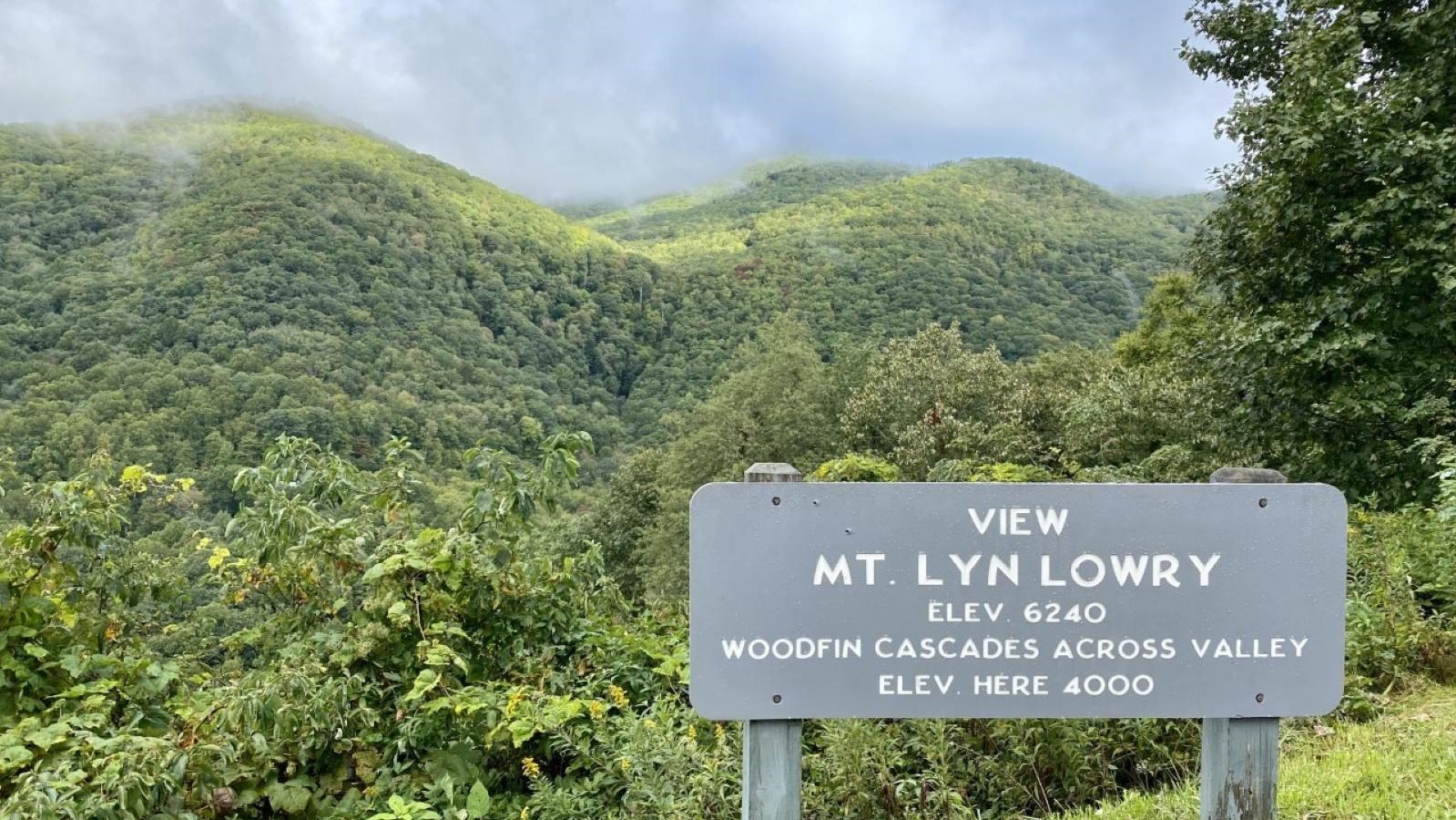 A gray overlook sign sits in front of green rounded mountains