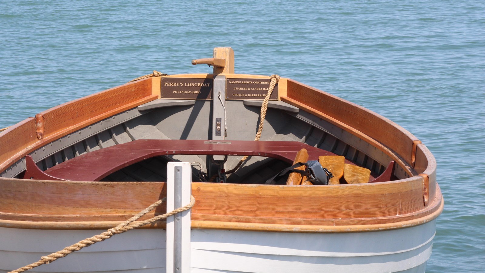 an 1812 replica of a white-painted, wood-lapped longboat with stained wood trim sits in a lake