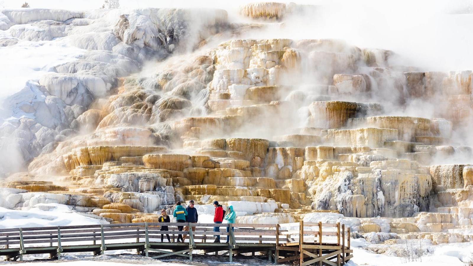 People stand on a boardwalk at the base of a steaming travertine terrace hydrothermal formation.