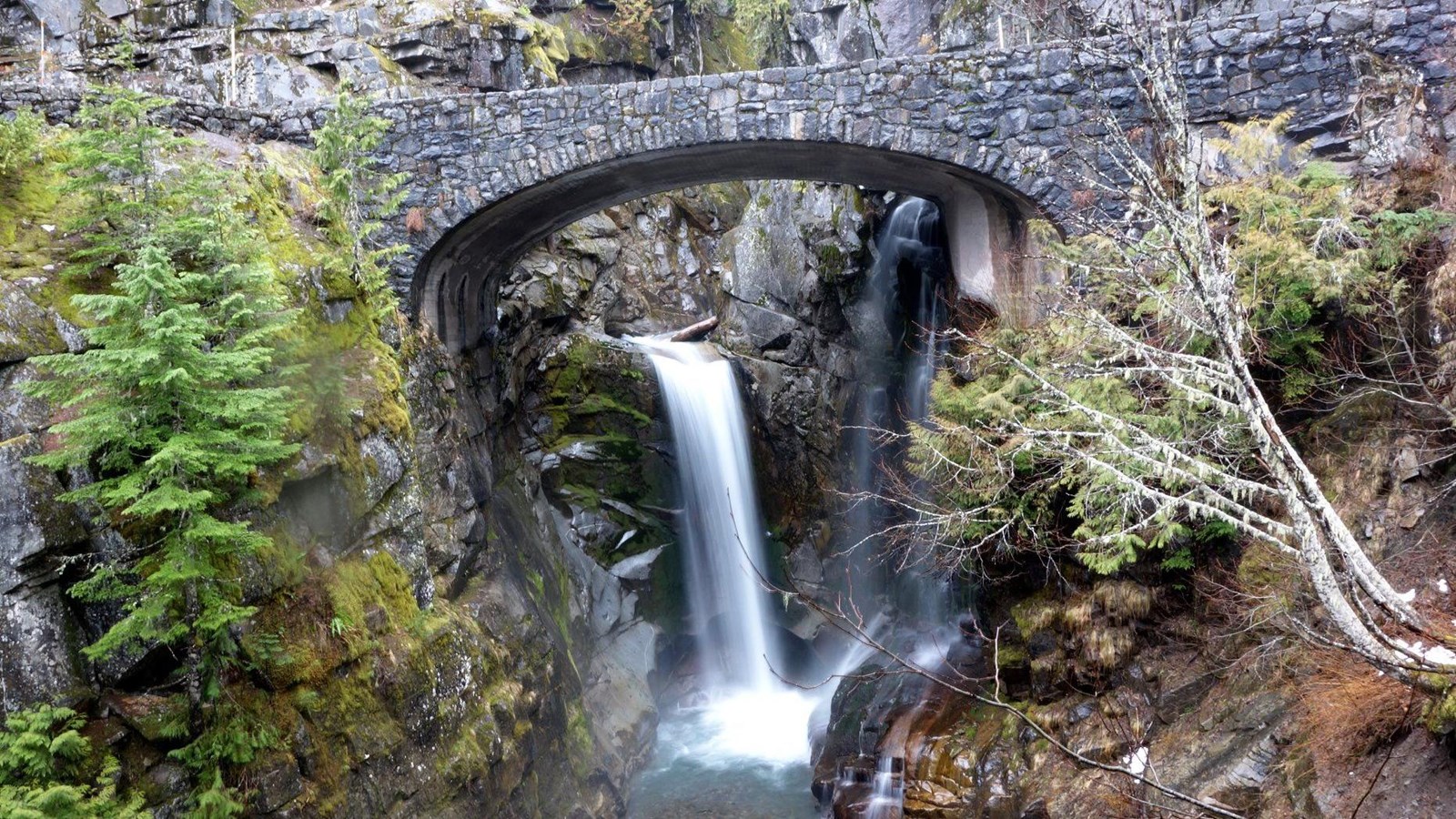 A gray stone bridge creates a unique frame for water cascading down a rocky ledge into a river below