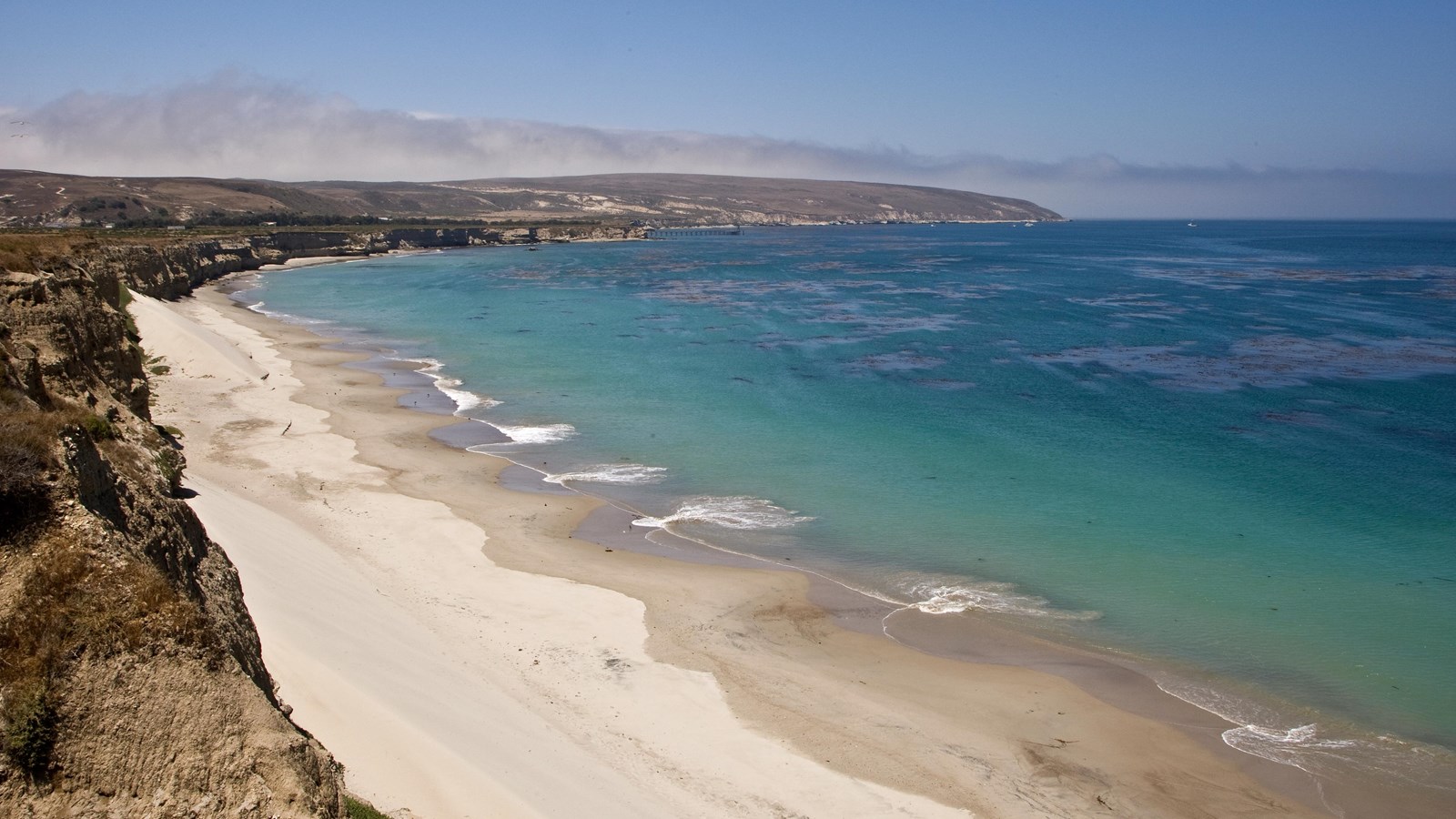 Coastal bluffs give way to white sand beach and aqua water with low hills in the background.