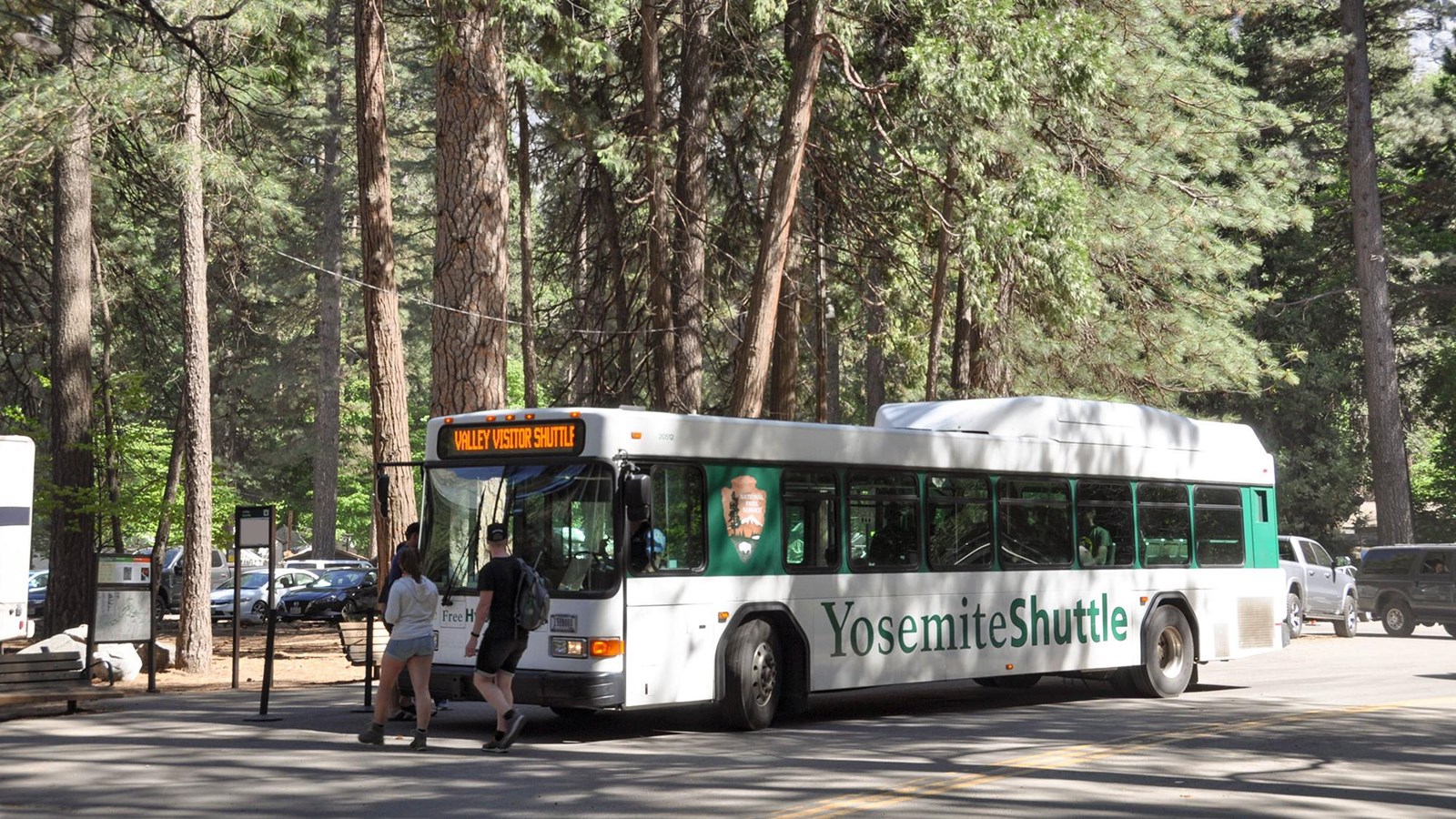 Shuttle at stop near dirt parking area with visitors crossing the street to get on the bus.