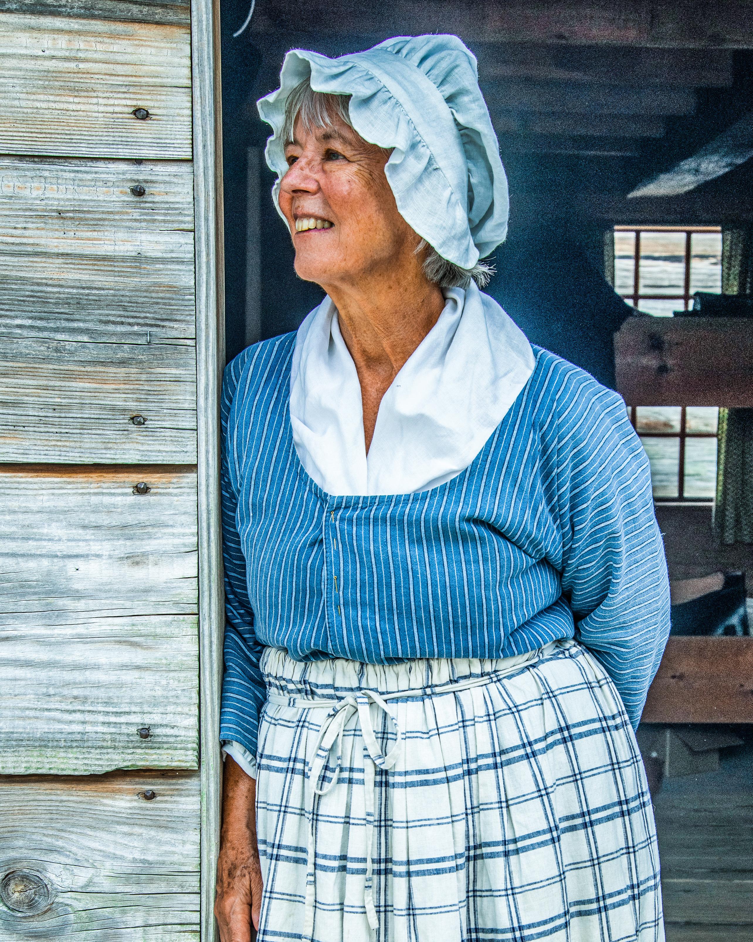A woman smiles, leaning on a door. A cap covers her hair, and the sun shines on her face. 