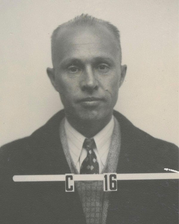 A black and white identification photo of a mostly bald man in a suit, with the number C16.