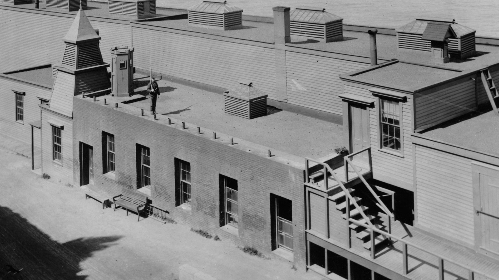 Black and white image looking down onto the lower prison building, a guard walks on the roof edge
