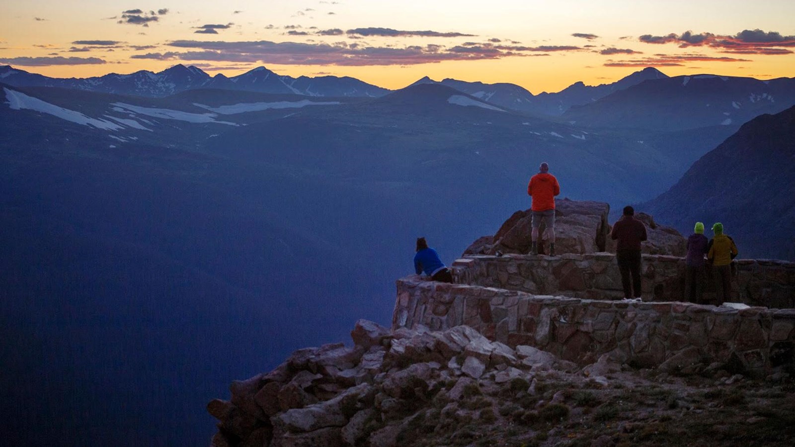 people stand on a platform watching the sun set behind mountains